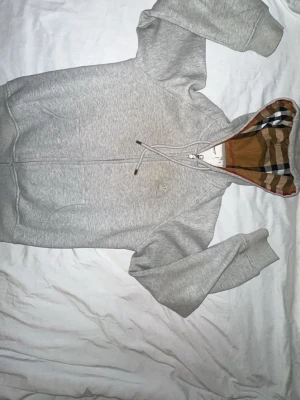 Grå zip-hoodie från Burberry - Snygg grå hoodie med dragkedja från Burberry. Klassisk huva med det ikoniska rutmönstret på insidan och dragsko. Broderad logga på bröstet och två fickor framtill. Perfekt för en stilren och avslappnad look.