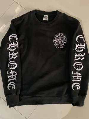 Svart Chrome Hearts sweatshirt - Svart sweatshirt från Chrome Hearts med tryckt logga på bröstet och stora CHROME-tryck längs båda ärmarna i vitt. Rund halsringning och ribbade muddar. Perfekt för dig som gillar streetwear och vill sticka ut med en ikonisk design.