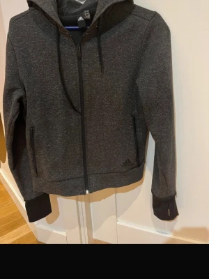 Mörkgrå hoodie från Adidas - Snygg mörkgrå hoodie från Adidas med dragkedja framtill och två fickor. Tröjan har huva med snörning och ribbade muddar vid ärmsluten. Perfekt för en avslappnad och sportig stil.