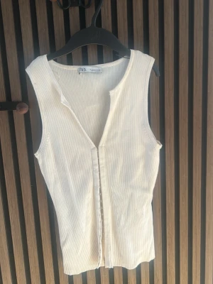 Vit ribbad ärmlös topp från Zara - Snygg vit ribbad topp från Zara med knappar framtill. Modellen är ärmlös och har en smickrande v-ringning. Perfekt att matcha med jeans eller kjol för en trendig look.