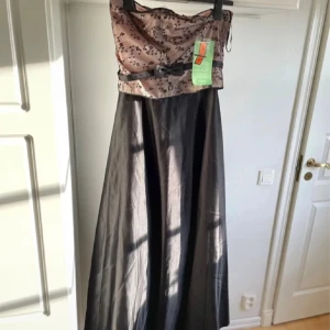 Långklänning med blommig topp och rosett - Balklänning. Elegant långklänning med blommig, ljusrosa topp och svart kjol. Klänningen är axelbandslös och har en dekorativ rosett i midjan. Perfekt för speciella tillfällen där du vill sticka ut med en klassisk och feminin look.