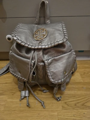 Silverfärgad ryggsäck  - Snygg ryggsäck i metallic silver med flätade kanter och logga på locket. Väskan har två framfickor med tryckknapp, justerbara axelband och dragskostängning. 