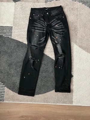 Purple Brand Byxor - Coola svarta jeans med slitningar på knäna och färgstänk i vitt och orange. Jeansen har en slim fit passform och klassiska fem fickor. Perfekt för dig som vill ha en edgy och trendig look.