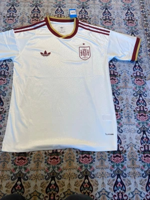 Vit Spanien tröja  - Säljer en vit Spanien tröja  med vinröda detaljer och klassiska Adidas-ränder på axlarna. Tröjan har Spanien-emblem på bröstet och Adidas logga. Materialet är lätt och andas, perfekt för sport och träning.