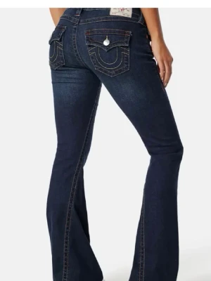 Mörkblå bootcut jeans från True Religion - Säljer ett par mörkblå bootcut jeans från True Religion (Joey modell) med ikoniska fickdetaljer och kontrastsömmar. Jeansen har låg midja, utsvängda ben och klassiska fickor med lock och knapp bak. Perfekta för en trendig och avslappnad look.