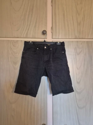 Svarta denimshorts med fem fickor - Säljer ett par svarta denimshorts med klassisk femficksdesign och knappgylf. Shortsen har raka ben och normal passform, perfekta för en avslappnad stil. Passar till det mesta och är ett måste i garderoben.    Alla kläder rollas innan dom skickas hem då det finns katt i hemmet🐈 Jag stryker och tvättar INTE innan jag skickar det får du stå för själv så det blir som du vill ha det🤗