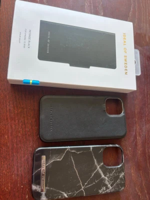 Två mobilskal från iDeal of Sweden - Säljer 3 snygga mobilskal från iDeal of Sweden. Ett är helsvart i läderlook och det andra har svart marmormönster med vita ådror. Båda har öppning för kamera och är designade för att skydda din mobil med stil. Perfekt för dig som vill ha ett trendigt och tåligt skal. Ett är helt oanvänt och är ett plånbokskal, med magnet så man kan ta av plånboksdelen. Inga sprickor på skalen. Passar till iPhone 12 Mini
