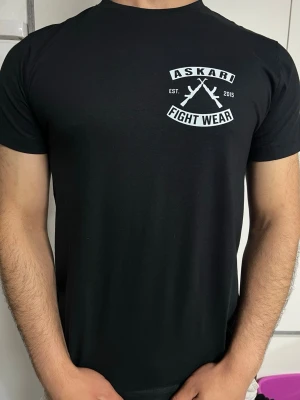 Svart Askari Fight Wear t-shirt - Snygg svart t-shirt från Askari Fight Wear med stort vitt tryck av korsade gevär och text både fram och bak. Klassisk rund hals och korta ärmar. Perfekt för dig som gillar streetwear och vill sticka ut med en bold design.