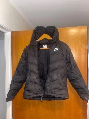 Svart pufferjacka från Nike - Säljer en svart pufferjacka från Nike med huva och dragkedja framtill. Jackan har två sidofickor och en vit Nike-logga på bröstet. Tillverkad i slitstarkt syntetmaterial som håller värmen under kalla dagar. Perfekt för dig som vill ha en stilren och sportig vinterjacka.