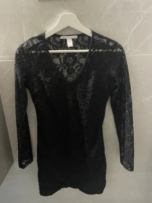 Svart spetsklänning  - Snygg svart kort spetsklänning från Rut&Circle med v-ringning och långa ärmar. Klänningen har ett genomgående spetsmönster som ger en elegant look. 