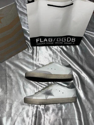 Golden Goose vita sneakers med glitter - Golden Goose sneakers i vitt läder med silverfärgad stjärna på sidan och glittrig häl. Klassisk låg modell med vita snören och grå sula. Märkesdetaljer på plös och häl. Perfekta för dig som gillar en unik och trendig look.24,5