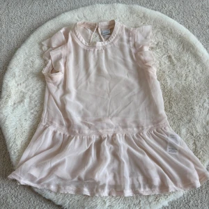 Ljusrosa volangblus från Gina Tricot - Söt ljusrosa blus från Gina Tricot med volangdetaljer på ärmar och nedtill. Blusen är ärmlös och har en flowy, luftig passform som ger en romantisk känsla.