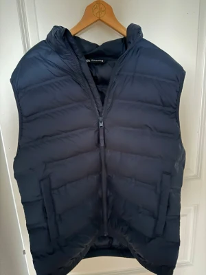 Svart dunväst från Zara - Snygg svart dunväst från Zara med dragkedja framtill och två stora fickor. Västen har en hög krage och quiltad design som ger en modern look. Perfekt att slänga över en hoodie eller tröja för extra värme. Materialet är mjukt och lätt, vilket gör den enkel att bära.