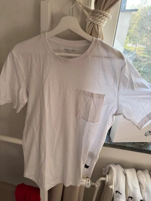 Vit t-shirt med bröstficka Jack & Jones - Klassisk vit t-shirt från Jack & Jones med rund halsringning och en enkel bröstficka på vänster sida. Tillverkad i mjuk bomull och har en normal passform. Perfekt basplagg för alla tillfällen.