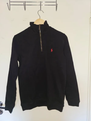 Svart stickad tröja Polo Ralph Lauren - Svart stickad tröja från Polo Ralph Lauren med halv dragkedja och röd broderad logga på bröstet. Hög krage och långa ärmar, perfekt för lager på lager. Klassisk och stilren design som funkar till det mesta.