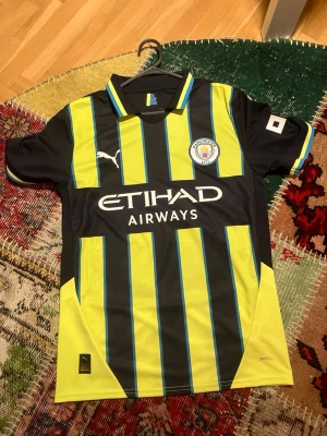 Manchester City bortatröja Puma - Snygg Manchester City bortatröja från Puma med svarta och gula vertikala ränder, blå detaljer och klubbmärke på bröstet. Tröjan har korta ärmar, krage och är tillverkad i ett lätt, ventilerande funktionsmaterial. Perfekt för fotbollsträning eller match.