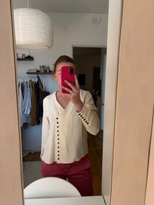 Asymmetrisk blus - Snygg krämvit blus med v-ringning och unika asymmetriska knappar längs framsidan. Långärmad modell med knappar även vid ärmsluten. Aldrig använd. 