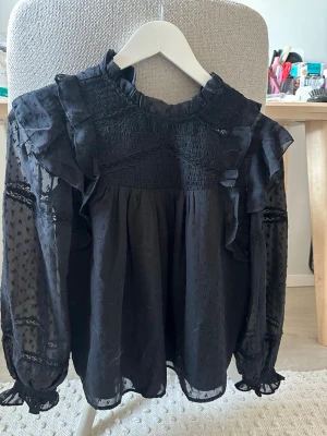 Svart volangblus med spetsdetaljer - Trendig svart blus med volanger och spetsdetaljer. Blusen har transparenta långärmade ärmar med små broderade prickar och resår vid ärmslut. Hög krage med volang och romantisk känsla. Perfekt för dig som gillar en chic och unik stil.