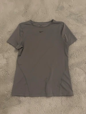 Grå Nike tränings t-shirt  - En stilren grå t-shirt från Nike med den klassiska svarta swoosh-loggan på bröstet. T-shirten har rund halsringning och korta ärmar. Tillverkad i ett mjukt och stretchigt funktionsmaterial som passar perfekt till träning. Använd ca 2 gånger