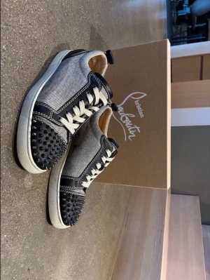 Christian Louboutin Louis Junior Spikes Svarta/grå sneakers - Christian Louboutin Louis Junior Spikes sneakers i svart och grått med vita snören och svarta dubbar framtill. Skorna har den klassiska röda sulan. Originalkvitto och dustbag medföljer. Storlek 41 1/2 men passar även 42 eller 41