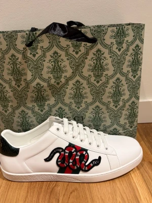 Gucci vita sneakers med orm -  vita sneakers från Gucci i läder med klassiska gröna och röda ränder på sidan och en broderad orm i svart, rött och vitt. Skorna har vita snören. Perfekta för dig som vill sticka ut med med en lyxig stil. 