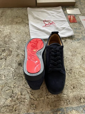 Mörkblå sneakers från Christian Louboutin - Snygga mörkblå sneakers från Christian Louboutin med klassisk röd sula och diskret logga. Skorna är låga och har ovandel i mocka med matchande mörkblå snörning. Insidan är fodrad i beige läder för extra komfort. Perfekt för dig som vill ha något exklusivt och stilrent.