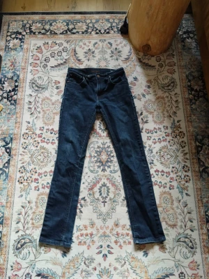 Mörkblå jeans från Nudie Jeans - Snygga mörkblå jeans från Nudie Jeans med klassisk femficksmodell och raka ben. Jeansen har en knappgylf och detaljer i metall. Tillverkade i slitstarkt denimtyg som ger en tidlös look.