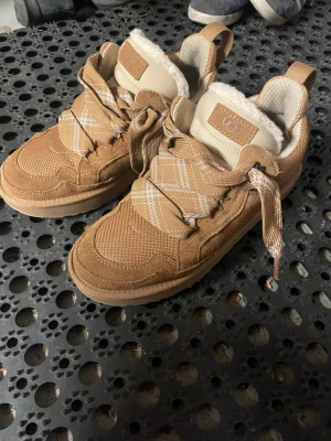 UGG Lo Lowmel - Säljer ett par snygga beige sneakers från UGG med breda rutiga snören och mysigt foder. Skorna har en kombination av mocka och textil på ovandelen samt en rejäl sula. Perfekta för dig som vill ha både stil och komfort.