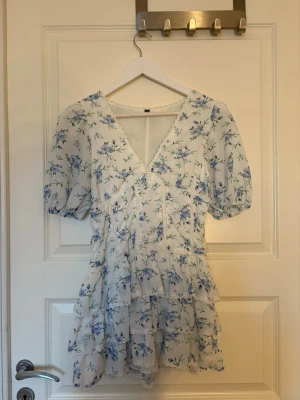 Vit blommig kortklänning med volang - Söt vit kortklänning med blå blommigt mönster och volangdetaljer. Klänningen har v-ringad omlottfront, puffade kortärmar och flera lager volanger. Perfekt för sommarens alla tillfällen. Materialet känns lätt och luftigt, troligen polyester eller bomull. Pris går att diskutera.