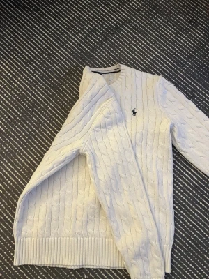Vit kabelstickad tröja från Polo Ralph Lauren - Säljer en klassisk vit kabelstickad tröja från Polo Ralph Lauren med rund halsringning och den ikoniska loggan broderad på bröstet. Tröjan har långa ärmar och ribbade muddar vid ärmslut och nederkant. Perfekt till alla outfits!