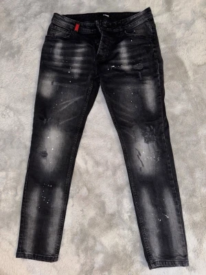 Svarta jeans med slitningar - Säljer ett par svarta jeans med coola slitningar och vita färgstänk. Jeansen har fem fickor, dragkedja och knappar framtill samt en röd detalj vid fickan. Materialet är stretchigt denim som sitter bekvämt. Lite liten i storlek så sitter tight om du har 32 har du mindre sitter den lite lösare.