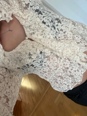 Vit spetsblus med volangdetaljer - Säljer en romantisk vit spetsblus med volangdetaljer längs kragen och knäppning framtill. Blusen är genombruten med blommigt spetsmönster och har långa ärmar. Perfekt för dig som gillar en feminin och trendig stil.