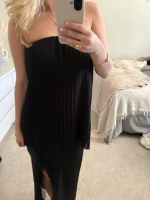 Svart plisserad tubtopp - Säljer en svart axelbandslös tubtopp med plisserad struktur. Toppen har en loose passform och är perfekt för dig som gillar stilrena och trendiga plagg. Materialet känns lätt och luftigt, vilket gör den skön att bära. Enkel att matcha med både kjol och byxor.