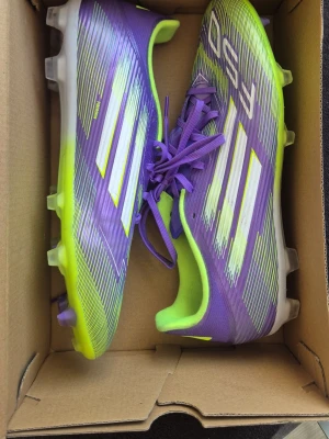 Lila Adidas F50 fotbollsskor - Säljer ett par Adidas F50 fotbollsskor i lila och neongult med vita detaljer. Skorna har klassisk snörning, syntetmaterial och mönstrad ovandel för bättre bollkontroll. Sulan har fasta dobbar i plast, perfekt för spel på gräsplan.