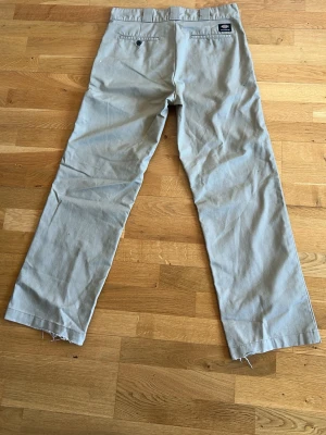 Beige chinos från Dickies - Säljer ett par klassiska beige chinos från Dickies med raka ben och avslappnad passform. Byxorna har fickor både fram och bak samt bälteshällor i midjan. Materialet är slitstarkt bomullstyg och byxorna har råa, avklippta benslut för en edgy look.
