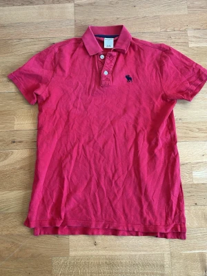 Röd pikétröja från Abercrombie & Fitch - Klassisk röd pikétröja från Abercrombie & Fitch med korta ärmar, krage och knappar framtill. Tröjan har en broderad mörkblå logga på bröstet och är tillverkad i mjuk bomull. Perfekt för en avslappnad och stilren look.