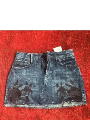 Blå jeanskjol med broderi från Guess - Snygg blå jeanskjol från Guess med broderade blommor framtill. Kjolen har klassisk femficksdesign, bälteshällor och rå kant nertill för en cool look. Perfekt för dig som gillar denim med en twist.