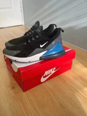 Nike Air Max 270 blå/grå/svart/vit - Nike Air Max 270 sneakers i svart mesh med grå och blå detaljer samt vit sula. Skorna har en stor synlig Air-enhet i hälen och klassisk Nike Swoosh på sidan. Lågt skaft och modern design som passar till streetwear. Materialet är en mix av mesh och syntet.