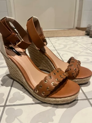 Bruna kilklackade sandaler från ONLY - Snygga bruna sandaler med kilklack från ONLY. Skorna har flätad sula i rep, bred rem framtill med guldfärgade öljetter och justerbart ankelband med spänne. Perfekta för sommaren. Helt nya aldrig andvända 