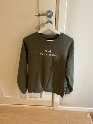 Grön sweatshirt från Peak Performance - Mörkgrön sweatshirt från Peak Performance med vit logotyptext på bröstet. Klassisk rund halsringning och långa ärmar. Tillverkad i mjukt material som passar perfekt för en avslappnad stil.