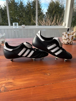 Adidas World Cup fotbollsskor svart/vit skruvdobb - Klassiska Adidas World Cup fotbollsskor i svart läder med vita ränder och skruvdobbar i metall. Skorna har låg profil, snörning och tunga med kardborre. Perfekta för spel på naturgräs och ger grymt grepp och kontroll på planen.