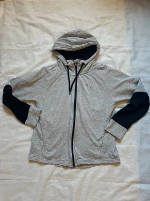 Grå zip hoodie från Puma - Snygg grå hoodie från Puma med dragkedja framtill och svarta detaljer på ärmar och huva. Hoodien har svarta snören och Puma-logga i silver på bröstet. Tillverkad i mjukt och bekvämt material, perfekt för en avslappnad stil.