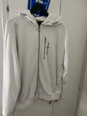 Vit hoodie från Sail Racing - Snygg vit hoodie från Sail Racing med dragkedja framtill och en bröstficka med zip och logga. Klassisk huva och ribbade muddar vid ärmslut och nederkant. Tillverkad i mjukt bomullsmaterial som känns skönt mot huden. Perfekt för en avslappnad och sportig stil.
