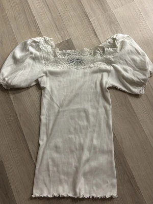 Vit topp med spets och puffärm - Supersöt vit topp med puffiga korta ärmar och spetsdetaljer vid halsringningen. Ribbad struktur i tyget och lätt volangkant nertill.  Aldrig använt och känner att den inte passar på mig och att den inte är i min stil 