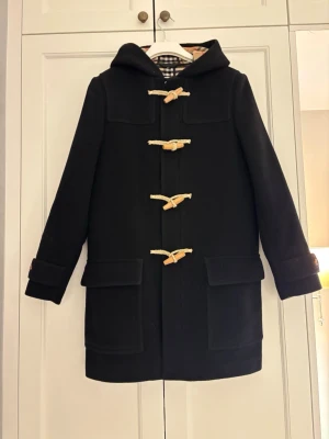Svart duffelcoat med träknappar från burburry - Klassisk svart duffelcoat med huva och träknappar i toggle-stil från Burberry. 