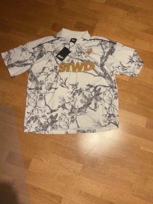Vit och grå mönstrad STWD t-shirt - Säljer en vit t-shirt från STWD med grått grenmönster och guldigt STWD-tryck på bröstet. T-shirten har korta ärmar, krage och är tillverkad i mjuk polyester. Perfekt för dig som vill sticka ut med en unik look. Ny oanvänd och riktigt bra style. Luftig så perfekt till varmt väder nu inför vår/sommar.