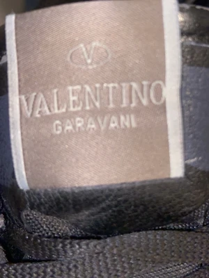 Svarta Valentino Garavani sneakers - Snygga svarta sneakers från Valentino Garavani med klassisk låg profil. Skorna har ovandel i textil och läderdetaljer samt svarta skosnören. Perfekta för dig som vill ha en exklusiv och stilren look. Använda ett par gånger, passar till alla årstider vatten täta också. STORLEK 42
