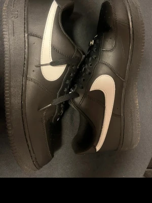 Nike Air Force 1 svarta sneakers - Svarta Nike Air Force 1 sneakers i läder med klassisk vit swoosh på sidan. Skorna har svarta skosnören, perforerad tåbox och låg profil. Tidlös streetstil som funkar till det mesta och ger en clean look.