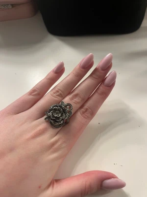 Stor silverfärgad blomring - Säljer en unik och iögonfallande ring i silverfärgad metall med en stor, detaljerad blomformad design. Ringen har fina snirkliga detaljer runt blomman och passar perfekt för dig som gillar statement-smycken.