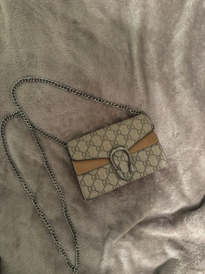 Gucci Dionysus axelväska beige - Snygg Gucci Dionysus axelväska i beige canvas med klassiskt GG-mönster och bruna läderdetaljer. Väskan har en silverfärgad kedjerem och ett dekorativt spänne framtill. Perfekt för dig som gillar lyxiga accessoarer och vill sticka ut.